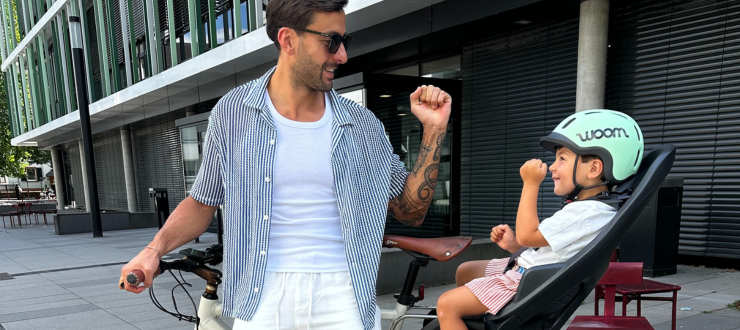 Ein Vater hält sein weißes Geero E-Bike. Er trägt eine Sonnenbrille und lässige Sommerkleidung, ein offenes weiß-blau gestreiftes Hemd und eine lockere weiße Hose. Auf dem Fahrrad ist ein Kindersitz am Sattelrohr montiert. Darauf sitzt ein kleiner Junge mit Fahrradhelm. Die beiden machen einen Handgruß mit ihren Fäusten und lächeln einander an.